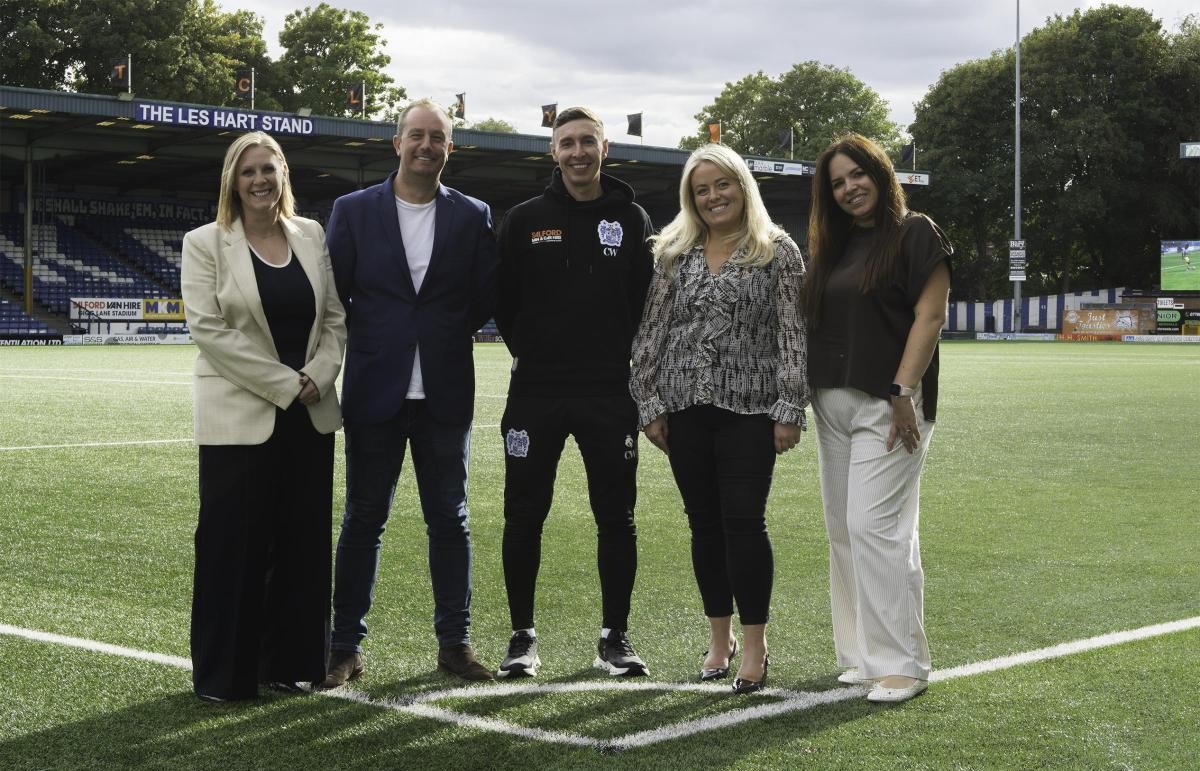 Members of the PM+M Bury team and a representative from Bury FC standing on the pitch at Gigg Lane to mark PM+M's tenth anniversary in Bury.